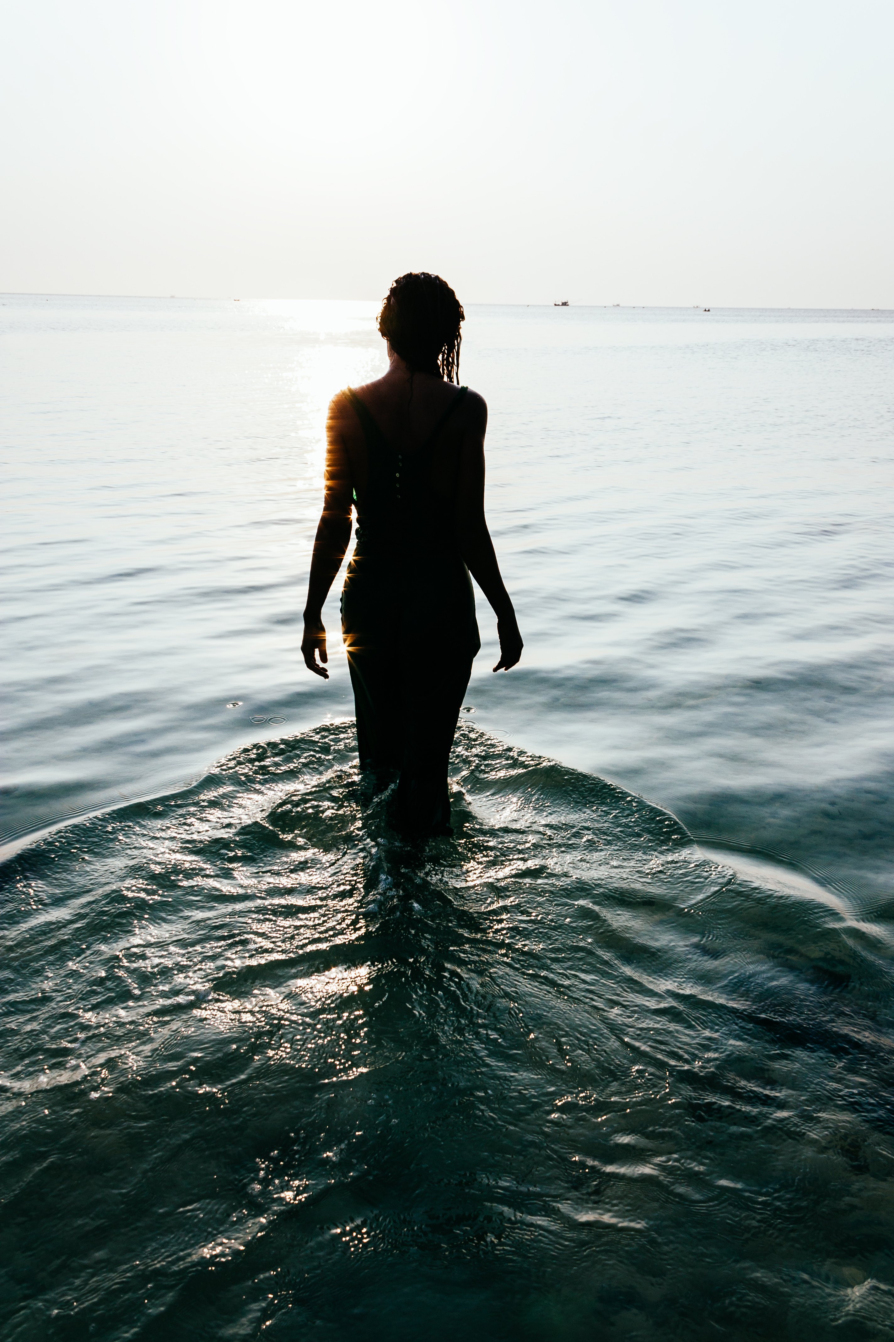 Tranquil scene with a view from behind of the silhouette of a wet young woman standing in the ocean facing out in meditation as the water flows around her legs creating a rippling pattern