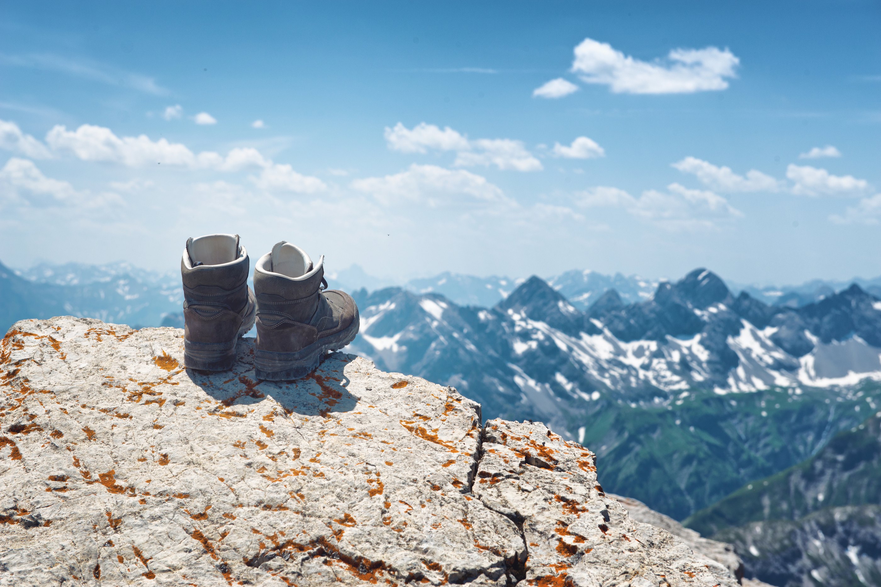 Pair of hiking boots on an alpine summit balanced on the edge of a rocky cliff overlooking distant alps with scattered snow on a sunny summer day, Hochvogel, Germany